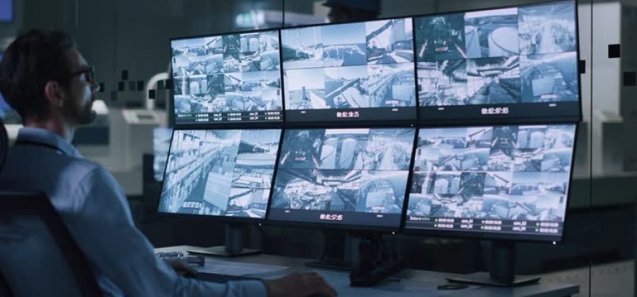 Industry 4.0 Modern Factory: Security Operator Controls Proper Functioning of Workshop Production Line, Uses Computer with Screens Showing Surveillance Camera Footage Feed. High-Tech Security